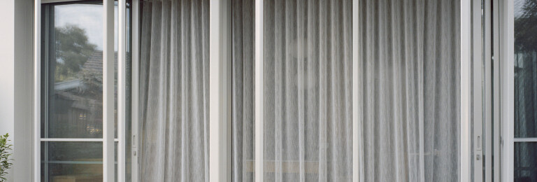 Sheer, dove grey curtains skirt the interior glazing on the ground floor, softening the transition between inside and outside.