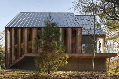 A slatted cedar screen spans the length of the west façade, concealing the entrance. At the front, an opening in the side wall allows access to a generous roof deck overlooking the lake.